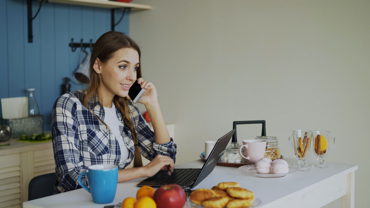 Woman at her kitchen table on the phone with a laptop, researching medication costs.