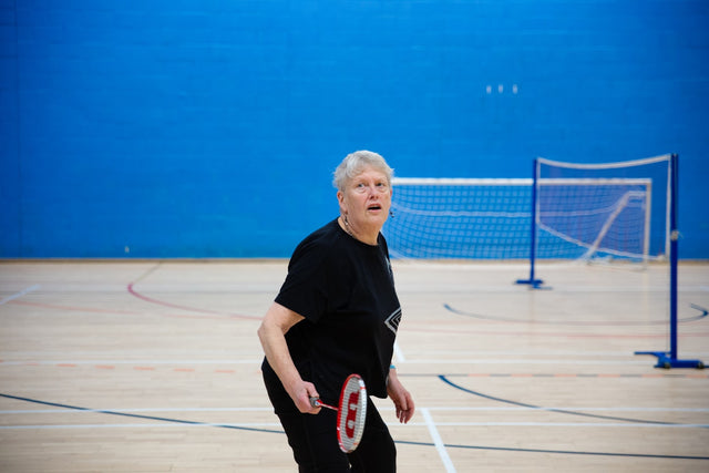 Elderly woman playing badminton in a gymnasium.