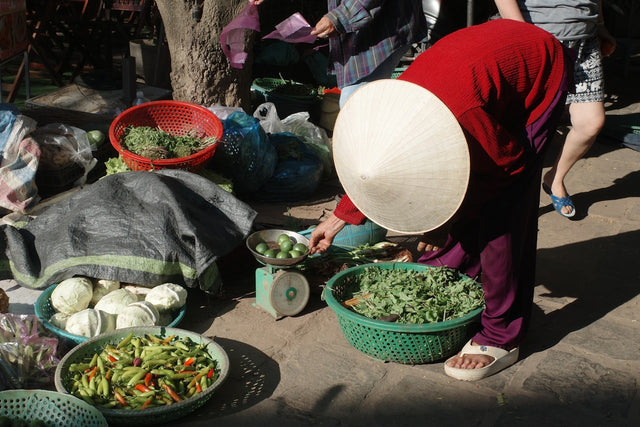 Vendor in conical hat sells fresh produce at a market.