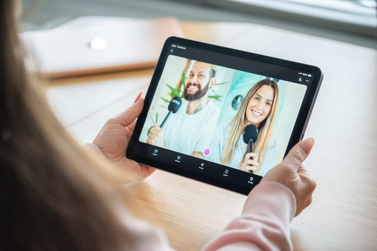 Person holds tablet displaying video call with two smiling hosts.